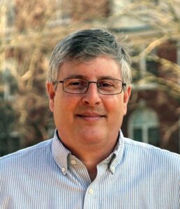 photo of a caucasian man with salt and pepper hair. he is smilng and wearing glasses. he is wearing a light blue button down shirt.