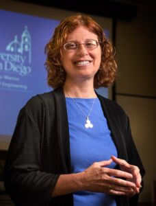 photo of smiling woman with shoulder length curly red hair. she is wearing glasses and wearing a blue top with black sweater. she is standing in front of a projected image that has math symbols